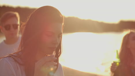 A-carefree-woman-student-is-dancing-on-the-open-air-party-with-with-beer.-She-smiles-and-touchs-her-long-dark-hair-and-enjoys-party-time-on-the-lake-coast-at-sunset.