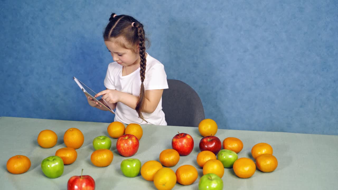 Little girl holding tablet in the kitchen. Sweet fruits laying on the table. Cute child playing games on gadget near the table with fresh apples and oranges.