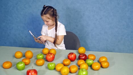 little girl holding tablet in the kitchen. sweet fruits laying on the table. cute child playing games on gadget near the table with fresh apples and oranges.