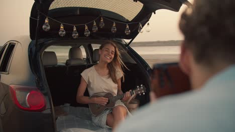 pov: a bearded man with curly hair in a blue shirt takes a photo of his blonde girlfriend, who is smiling and playing the ukulele in the trunk of a black car decorated with lights and shows her the photo against a yellow sky