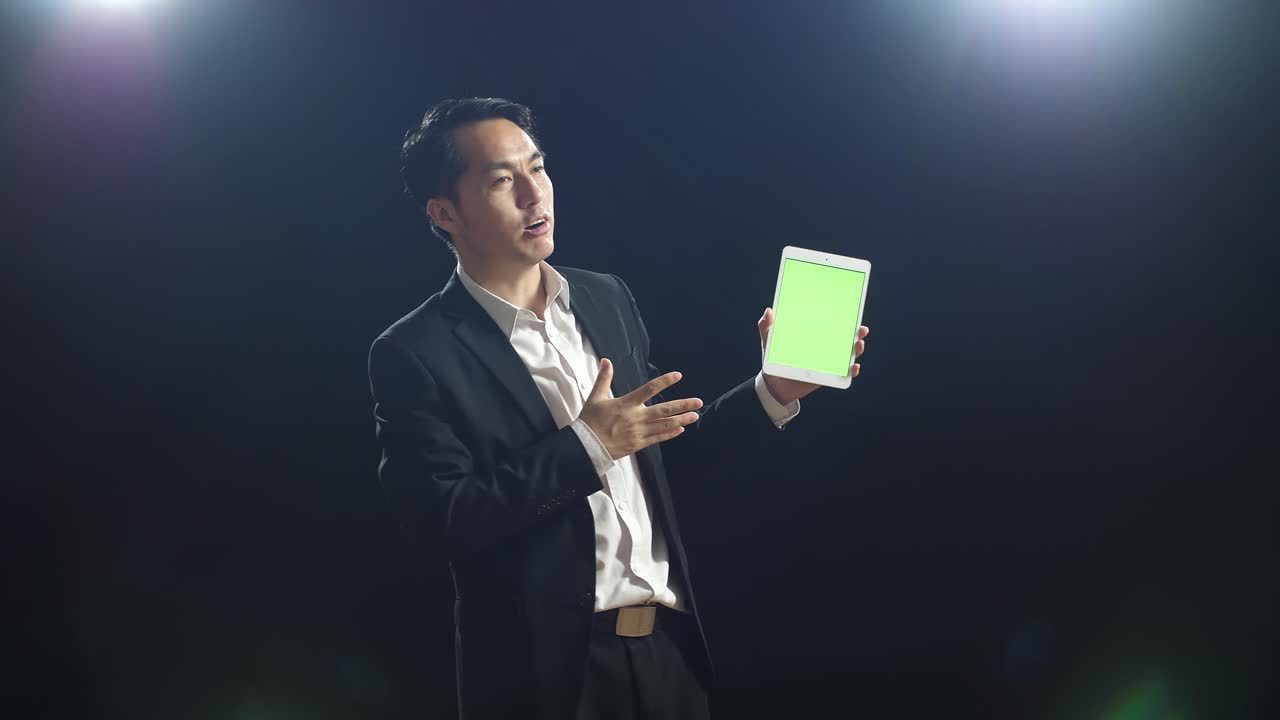 ブラックスクリーンスタジオで話している時ビジネススーツを着たアジア人スピーカーが緑色のスクリーンタブレットを握り指差しています