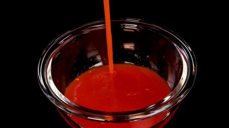 Un pomodoro maturo viene fatto cadere in una ciotola di vetro trasparente piena di salsa rossa, creando uno schizzo drammatico. Illuminazione da studio, sfondo nero, slow motion