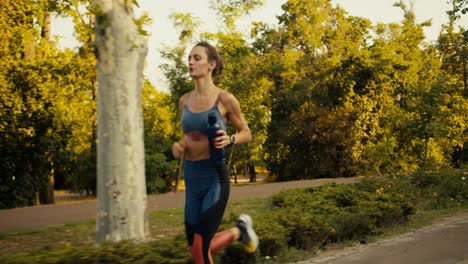 una chica deportiva en un uniforme de verano deportivo corre en un parque soleado por la mañana junto con una botella negra de agua