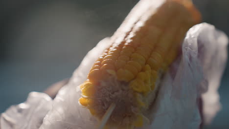 close up of corn being rotated with steam rising, person holding corn with plastic wrap, soft light illuminating kernels, blurred background with distant figures and warm outdoors