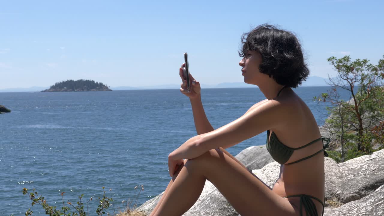 Girl in bikini sitting on rock at beach filming scenery with smartphone