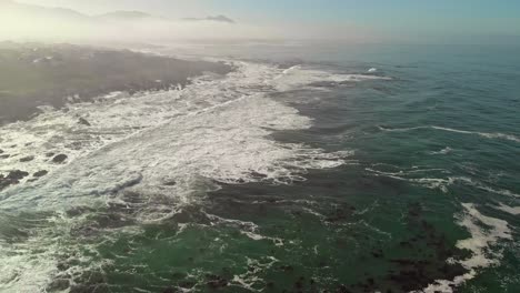 Vista-Aérea-De-Alto-Vuelo-Del-Océano-Y-La-Tierra-Mientras-Las-Olas-Llegan-Mientras-Se-Elevan-Desde-Las-Aguas-Para-Mostrar-La-Costa-Con-Un-Poco-De-Niebla