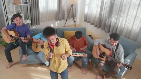 group of young asian friends playing rock music, a boy with headphones holding a microphone and singing while his friends playing musical instruments on a sofa at home