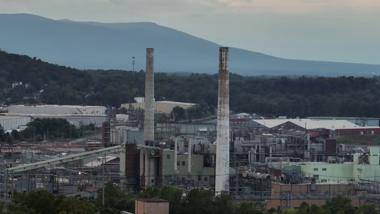 Aerial zoom of two smokestacks on industrial factory power plant in waynesboro. Virginia. Dark cloudy day in suburbia. Blue ridges mountains in background. Pipelines connecting storage
