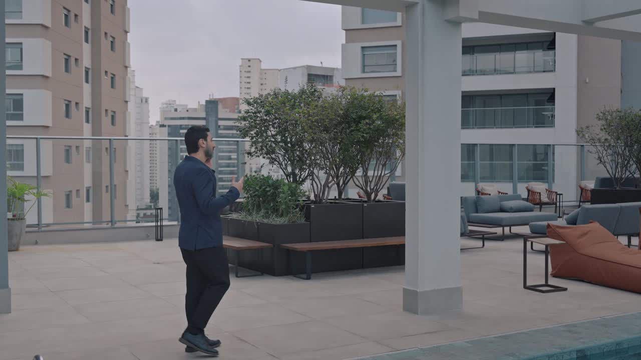 Executive man talking on the phone while walking along the rooftop terrace of a modern building. Pan right following the man.