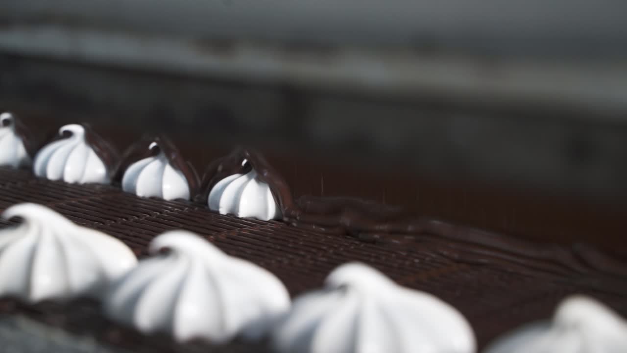 A white sweet marshmallow on a ribbon is covered with melted hot dark chocolate. Close-up. Conveyor at a confectionery factory