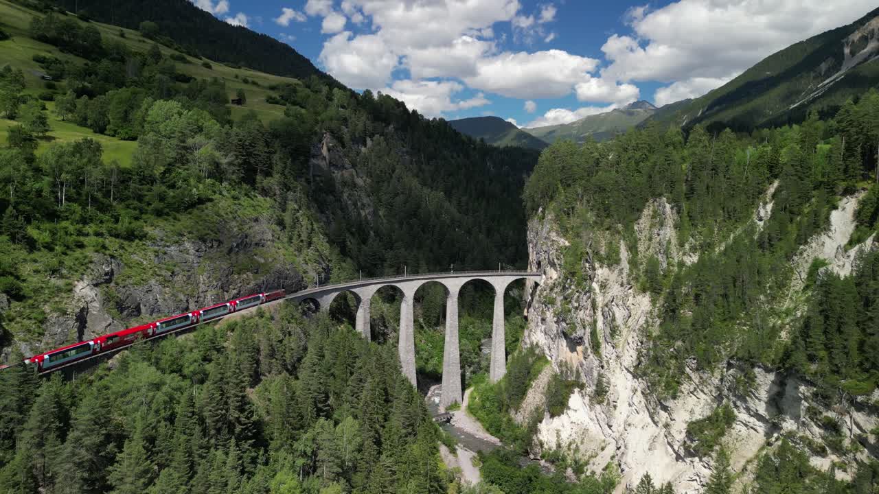 Train crossing Landwasser Viaduct between swiss alps entering tunnels in mountains covered in vibrant green trees and pure tranquil nature on sunny summer day