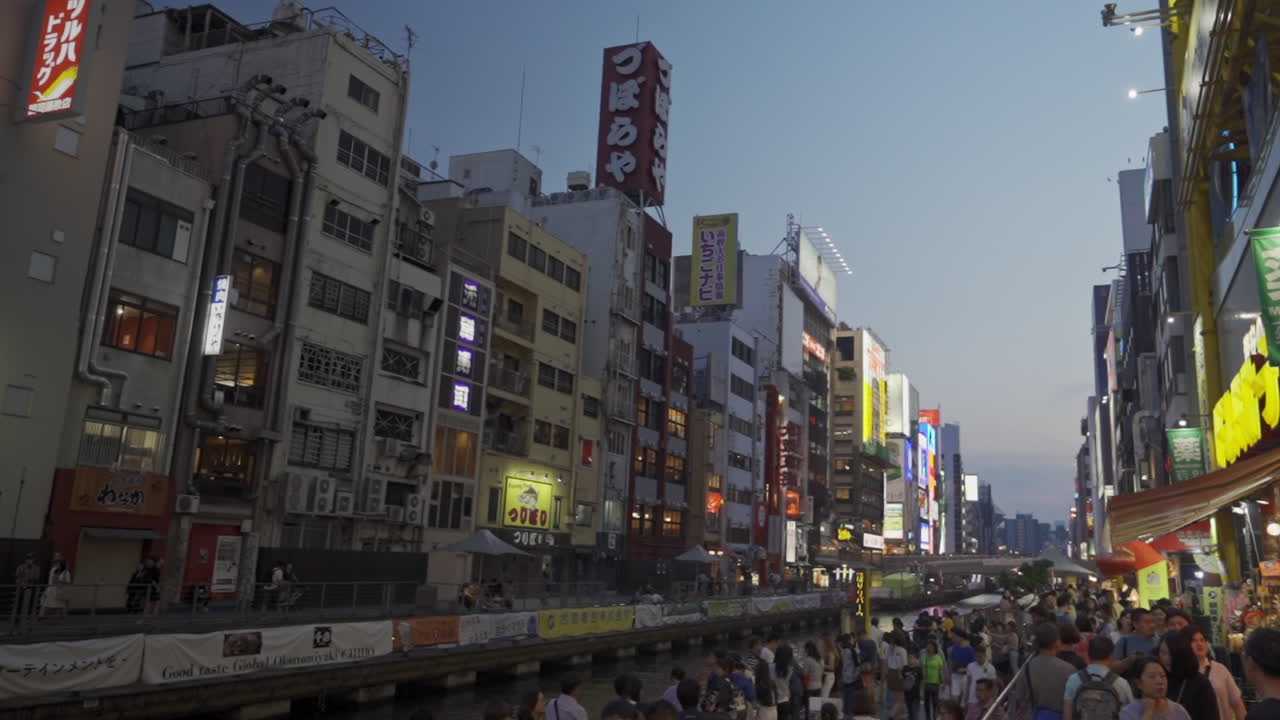 Along the Dotonbori canal with signs illuminated at dusk, Osaka, Japan, wide dolly in slow motion.