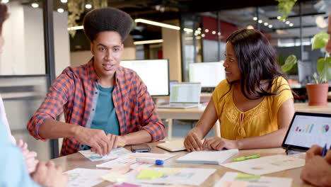 happy diverse colleagues brainstorming with documents and tablet at table in office in slow motion