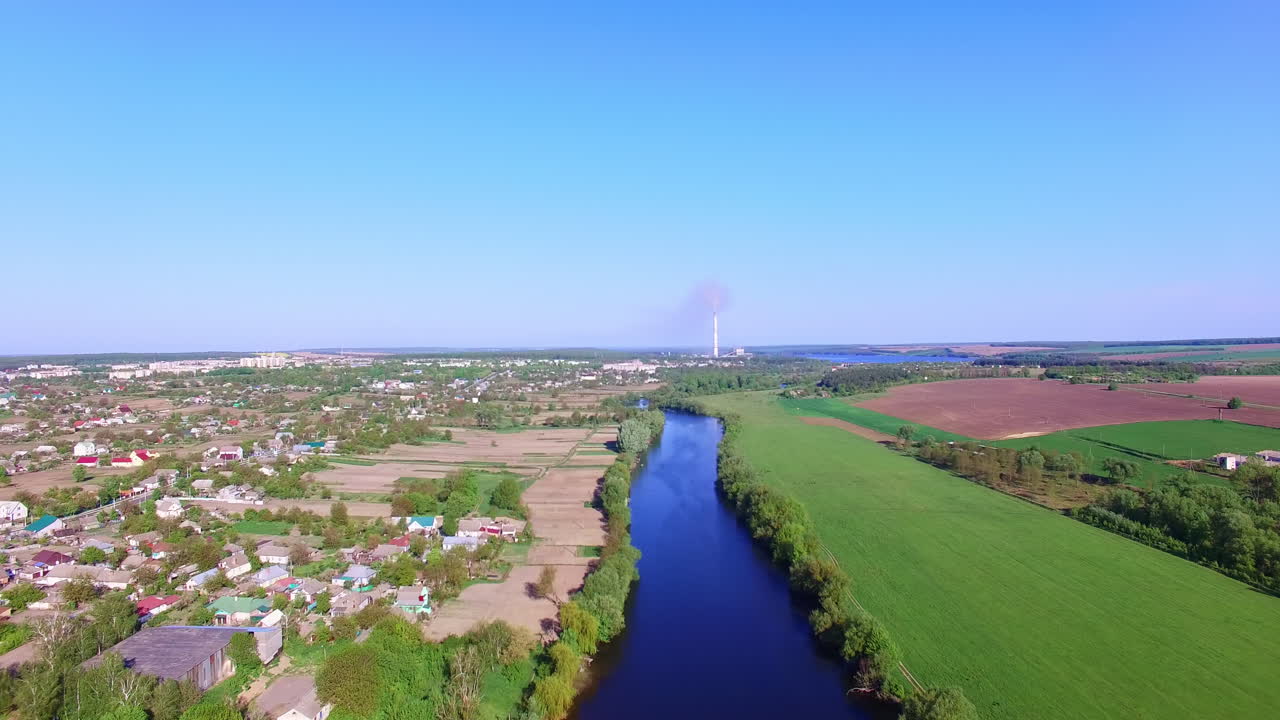 Large village with private houses outlined by thin blue river. Rural farmlands on the other side of the waterway. Top view.