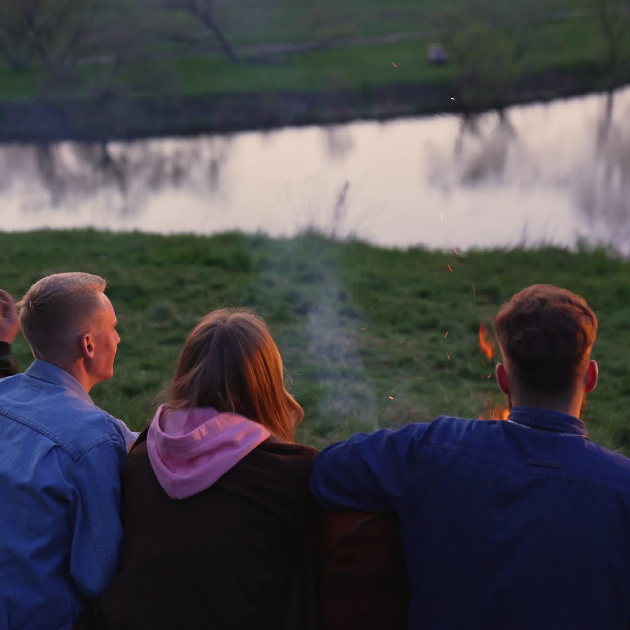 Group of young people sitting on the grass hear campfire and singing songs. Friends enjoy the sunset and view of the river in front of them