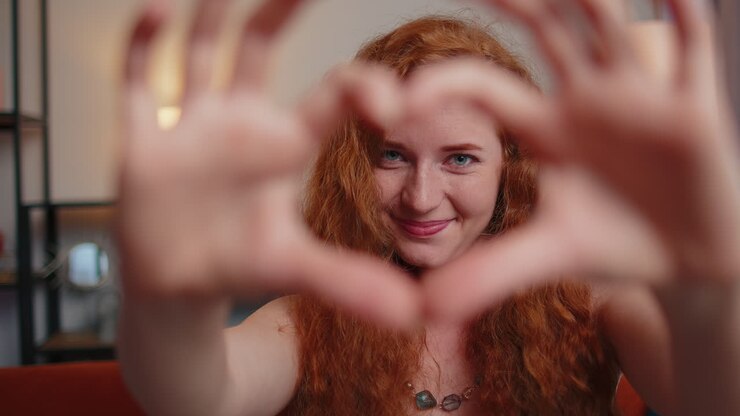 Giovane donna dai capelli rossi crea un simbolo d'amore, mostrando il simbolo del cuore alla fotocamera, esprimendo sentimenti romantici