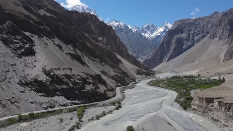 Hunza-Valley,-Pakistan,-Aerial-View-of-Scenic-Route-by-Glacial-River-Water-Under-Snow-Capped-Mountain-Peaks