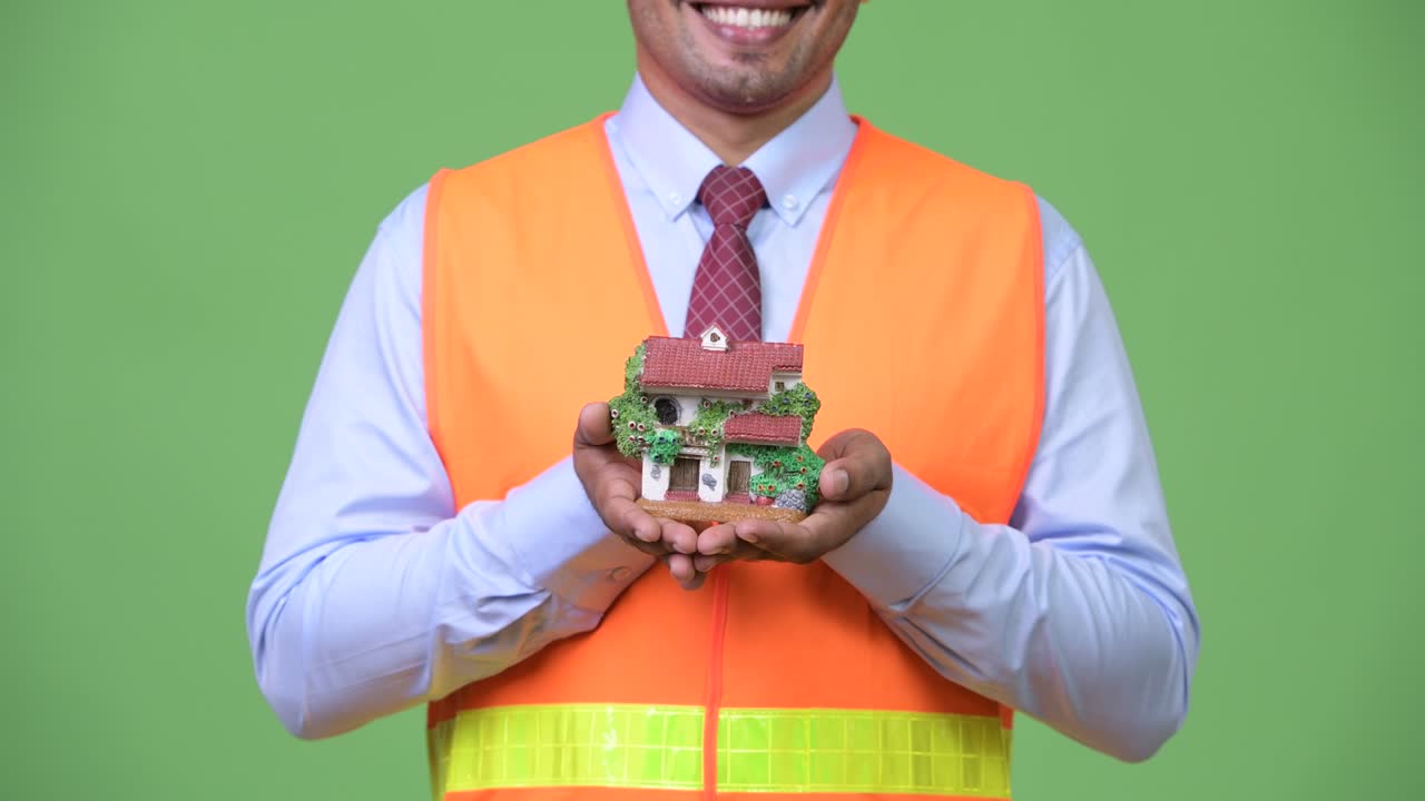 Young handsome Persian man construction worker holding house figurine