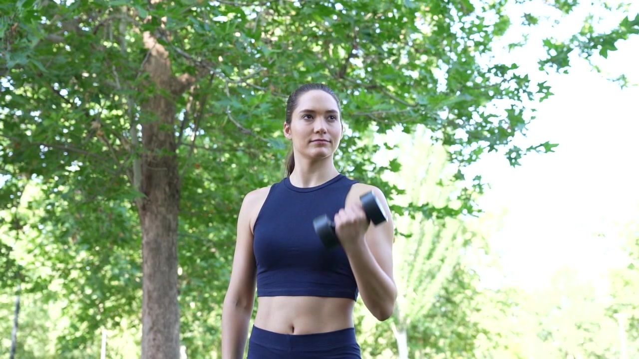 Joven mujer atlética haciendo ejercicio con mancuernas en un parque durante una sesión de entrenamiento al aire libre.