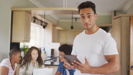 Feliz-Hombre-Latino-De-Pie-En-La-Cocina-Con-Tablet-Y-Diversos-Amigos-En-Segundo-Plano.