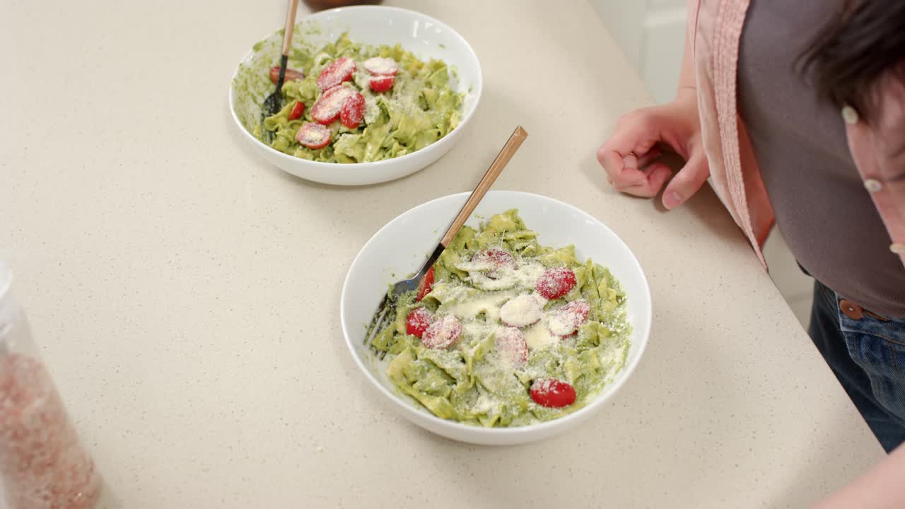 Person is shifting right hand after leaning, steadying two white bowls of green pasta at countertop