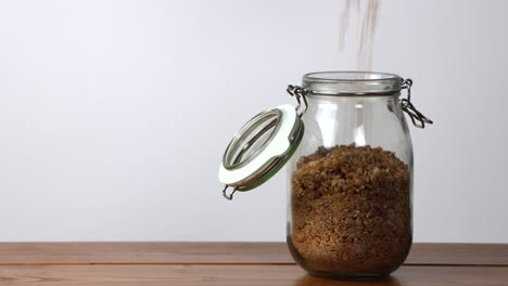 Cereal-Que-Se-Vierte-En-Un-Frasco-De-Vidrio-Sobre-Una-Mesa-De-Madera-Con-Fondo-Blanco