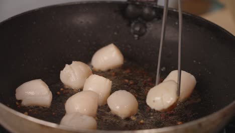 Delicious-seafood-dish-cooking-in-progress,-profession-chef-using-precision-tongs-to-flip-side,-making-sure-each-scallops-are-sauteing-evenly-with-the-garlic-on-the-frying-pan,-close-up-shot