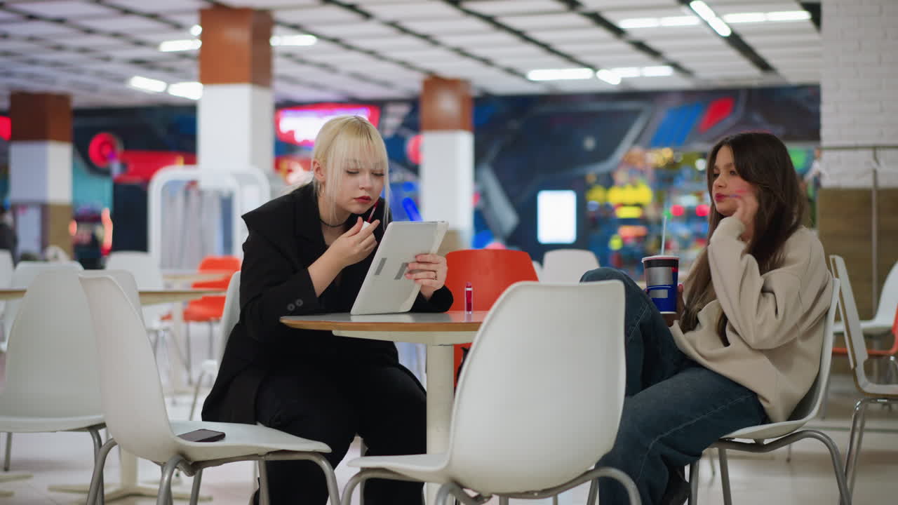 Two women sitting in bright cafe as one carefully applies lipstick using tablet mirror while other adjusts hair sipping drink, showing casual friendship moment with modern indoor background
