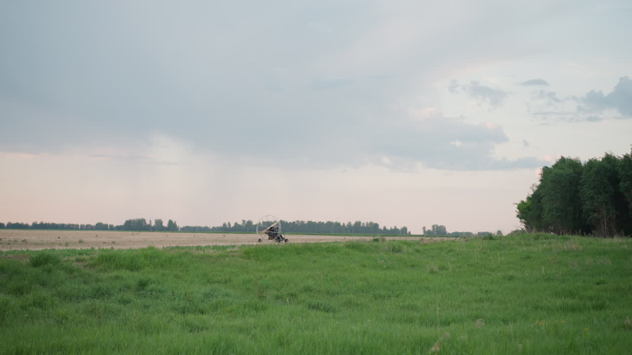 pilot seated in paramotor trike rolling across grassy field under cloudy sky near distant tree line preparing for takeoff with powered paraglider fan spinning on metal frame in tranquil rural setting