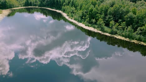 Die-Luftaufnahme-Fängt-Die-Abstrakte-Spiegelung-Eines-Bewölkten-Blauen-Himmels-In-Einem-Ruhigen-See-Ein,-Der-Von-Dichtem-Grünen-Wald-Umgeben-Ist