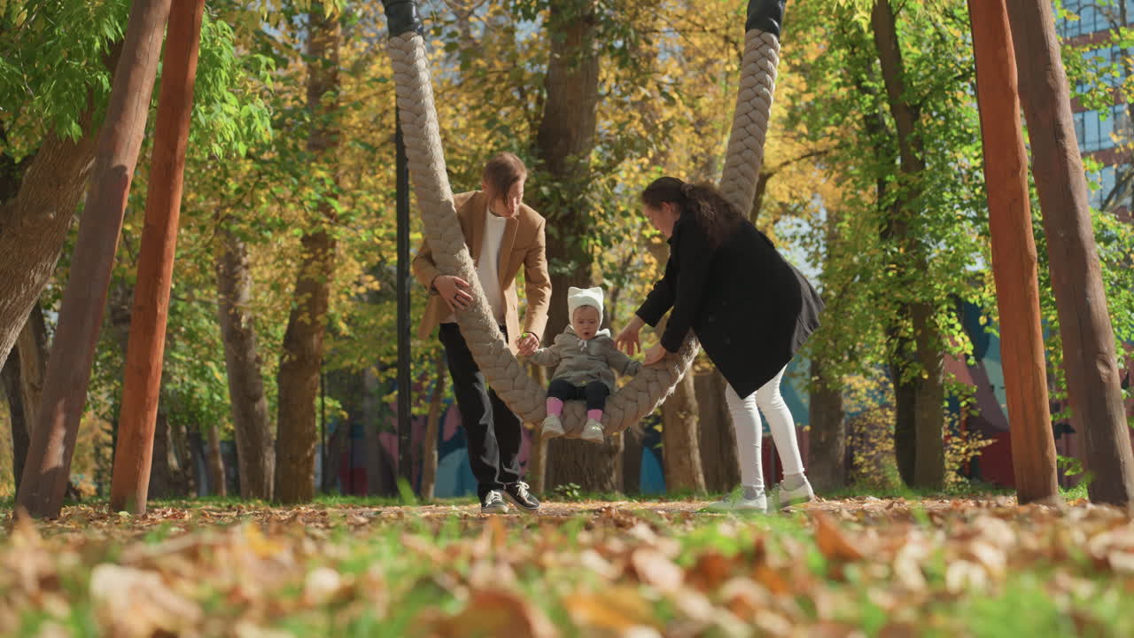 Niño pequeño en un columpio en el parque. Madre blanca y padre negro columpian suavemente al niño. Hojas de otoño cayendo, luz solar a través de los árboles. Pareja casada compartiendo un momento lúdico, con un vínculo estrecho y risas, manos.