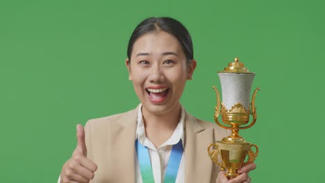 close up of asian business woman in a suit with a gold medal and trophy showing thumbs up gesture and smiling to camera as the first winner on green screen background in the studio