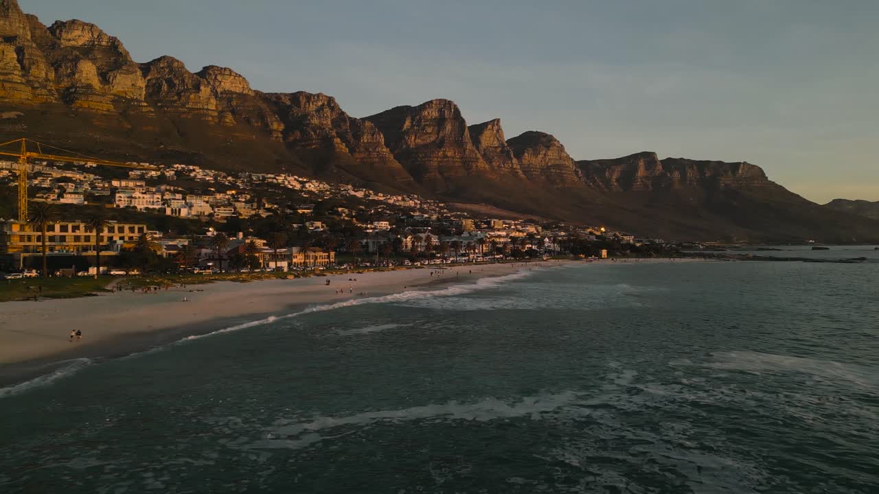 Plynulé letecké záběry z dronu v nízké výšce letící směrem k Camps Bay v Kapském Městě v Jižní Africe při západu slunce, zachycující zlaté světlo nad pobřežím.
