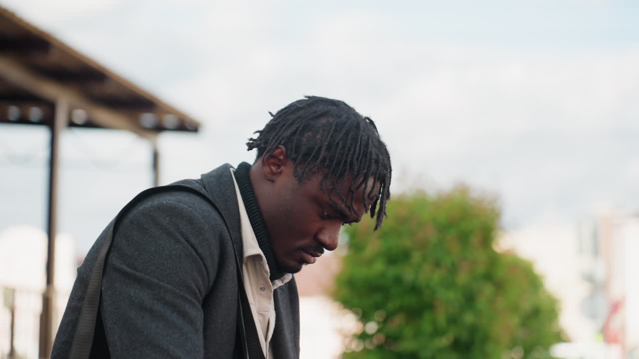 Close-up portret van jonge man met locs onder een doffe bewolkte hemel, met een gefocuste, bedachtzame uitdrukking en natuurlijke emotie, met zachte achtergrondvervaging die de kalme sfeer en de buitenomgeving benadrukt