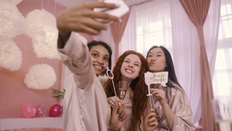 tres amigos multiétnicos con decoración de cartón y copas de champán de cristal tomando un selfie con smartphone en despedida de soltera