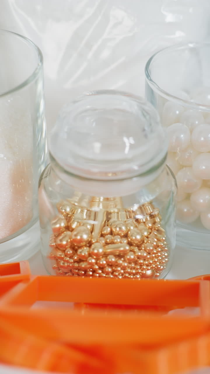 유리 항아리, 유리 컵에 설탕, 주방 카운터에 색 그에 놓인 오렌지색 베이킹 도구, 반이는 식용 진주 및 설탕 결정에 있는 다채로운 장식 된 구슬의 클로즈업