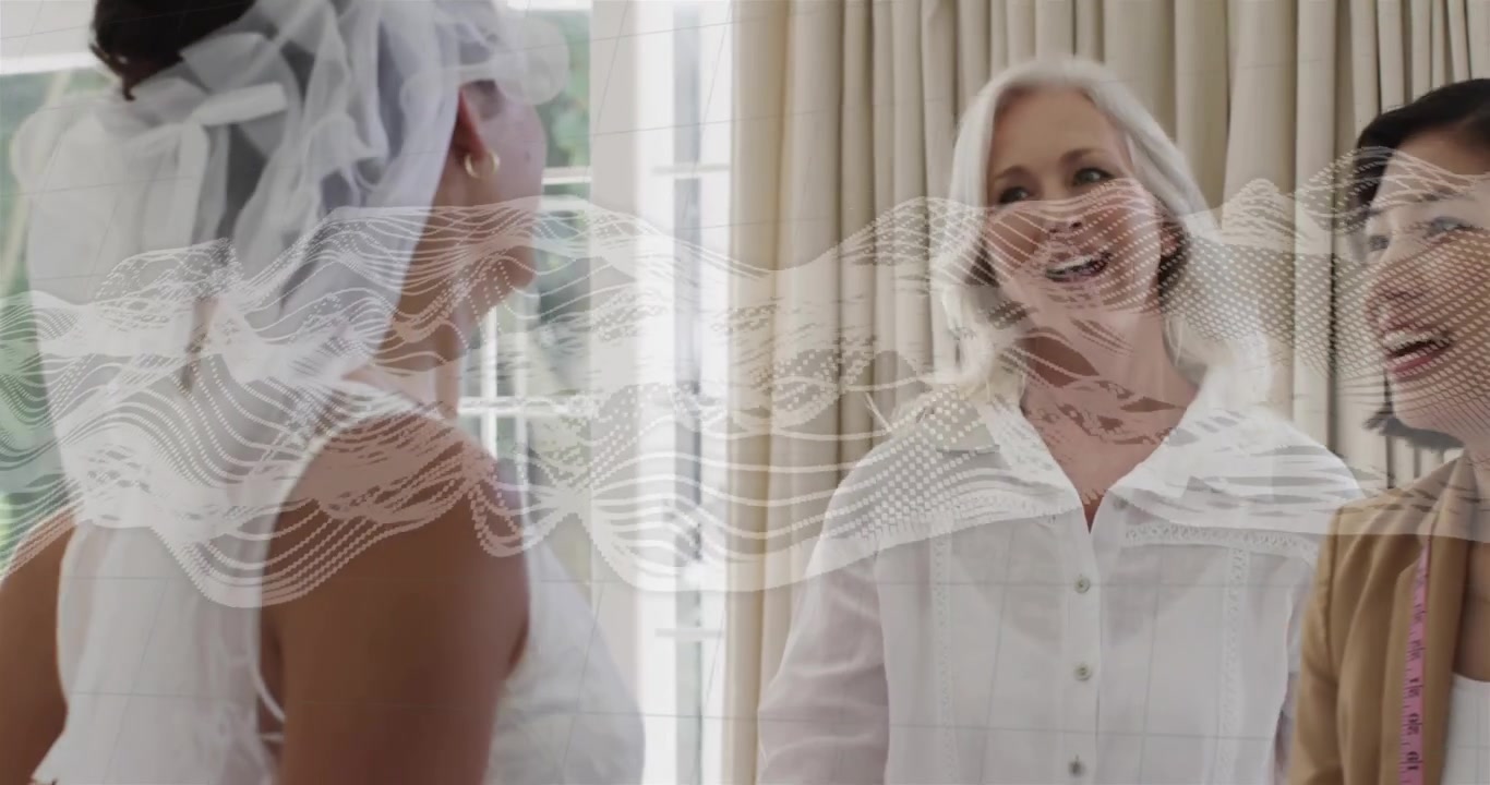 Sposa in preparazione nuziale che solleva il velo vicino alla finestra, mentre l'assistente inizia ad appuntare e la madre approva.