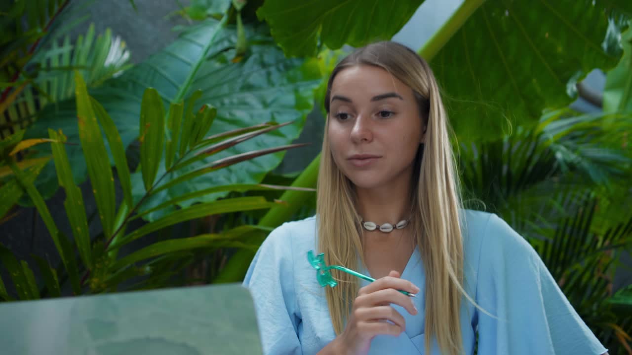 A girl in a blue dress remotely online working behind a laptop and looking into the screen the backyard with green plants