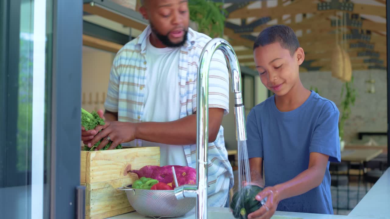 African American father and son rinsing produce at sink with tall faucet running, unpacking crate