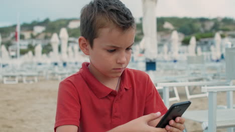 Chico-Serio-Sosteniendo-Un-Teléfono-Móvil-En-La-Playa.-Chico-Mirando-Al-Teléfono-Celular-En-La-Playa.