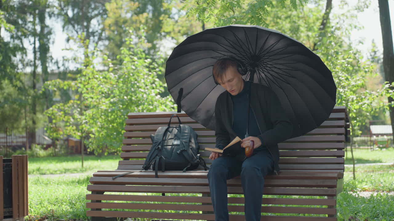 Uomo caucasico che legge sotto un ombrello su una panchina del parco, pausa riflessiva in un parco illuminato dal sole, ombrello di pizzo nero che protegge dalla luce, borsa di pelle accanto a lui, dita che sfogliano le pagine di un romanzo consumato