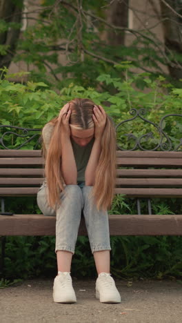 mujer se sienta en un banco de madera, descansando la cabeza en las manos en una postura pensativa y sombría, rodeada de vegetación y un edificio urbano en el fondo. su largo cabello fluye hacia abajo mientras ajusta su cabeza