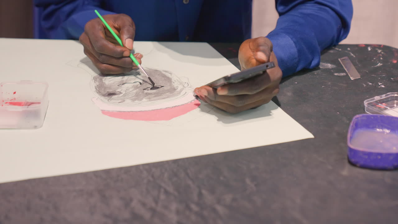 Black artist finishing portrait with fine detail, darkskinned male using small green brush to refine black ink textures on white paper, phone reference held in left hand, palette container at right