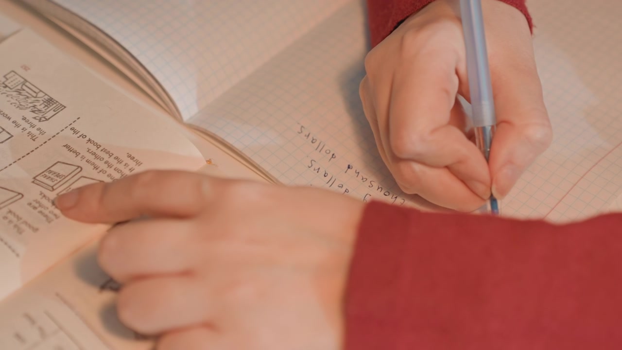 Close up of hands writing on graph paper. Notebook contains handwritten notes about money. Arm wearing red fabric is visible throughout scene interacting showing hand pen