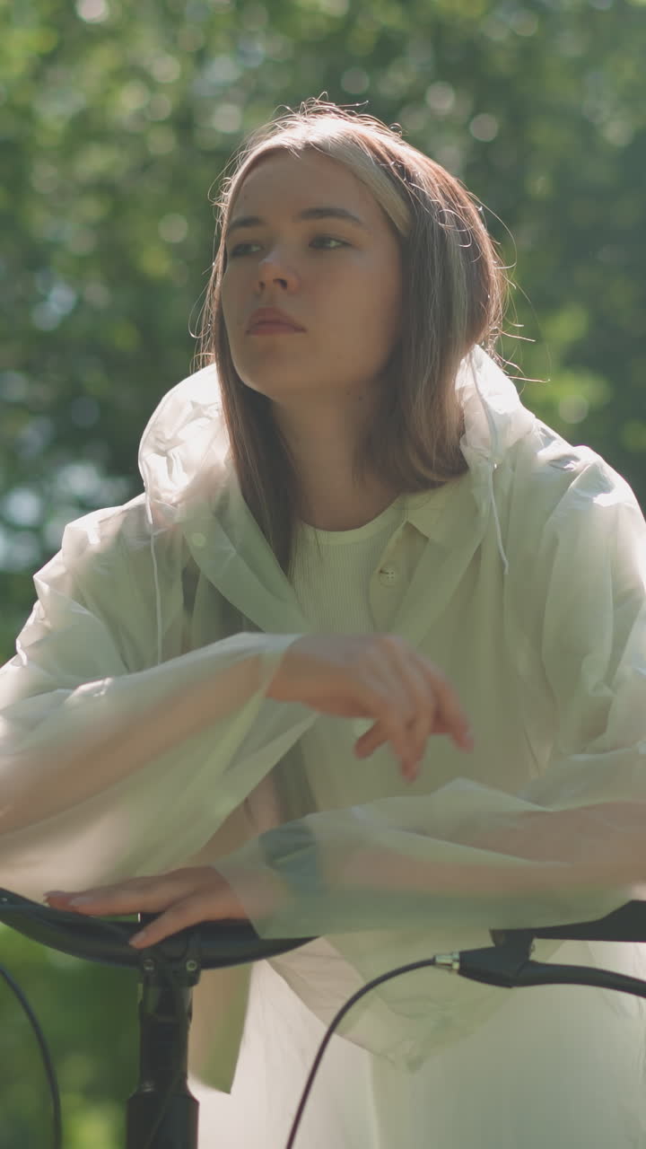 una mujer joven con un impermeable translúcido se quita la capucha, se apoya pensativamente en el manillar de la bicicleta con la mano apoyada en la barbilla, la luz del sol se filtra suavemente a través de las hojas de los árboles