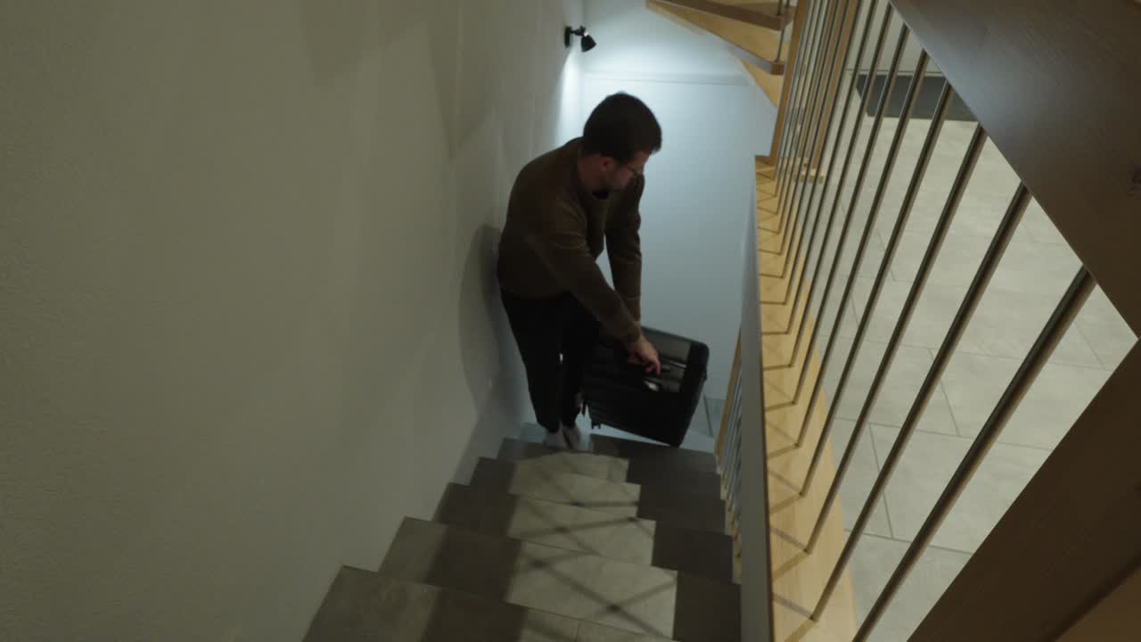 A man lifts and carries a heavy suitcase up a staircase, showing physical effort, travel preparation, exhaustion, and the challenge of moving luggage through narrow stairways