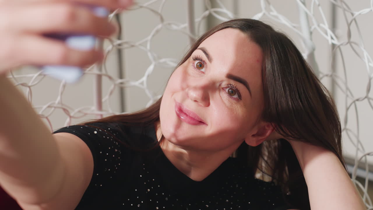 Selfi divertida de una mujer caucásica que se arregla el pelo con el smartphone, sonriendo y recogiendo su cabello mientras posa sentada en una silla colgante, con ropa casual, iluminación suave en casa y un ambiente desenfadado de redes sociales