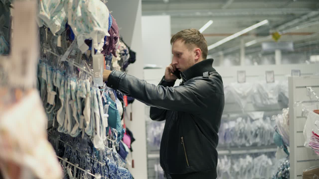 Young man is trying to choose a bra in lingerie store for his girlfriend or wife while talking on the phone and asking about size and color