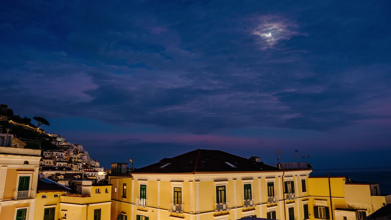 Timelapse del tramonto sopra i tetti della città di Amalfi con nuvole rosa tenui e orizzonte marino calmo in un'incantevole città costiera mediterranea. Timelapse dal giorno alla notte di un viaggio attraverso una storica città italiana
