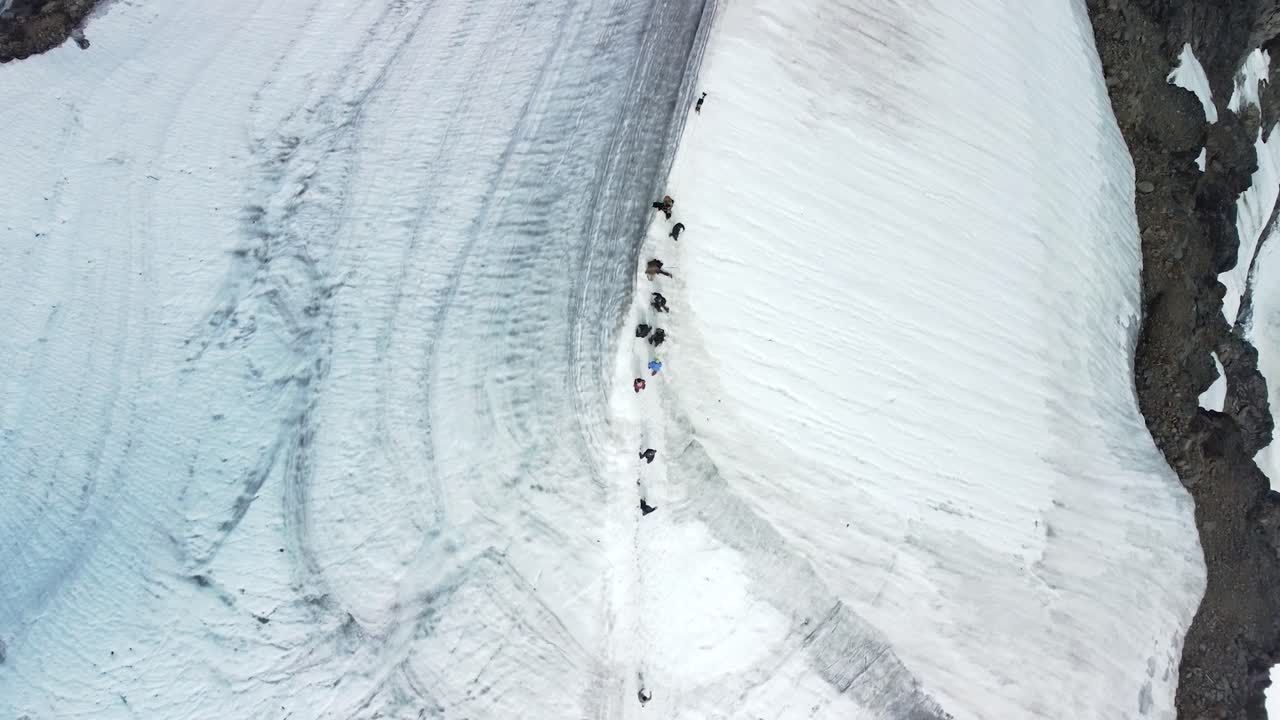 Top down aerial drone footage flying over a group of people walking up a white snow covered Kebnekaise mountain ridge during a cloudy day. People have colorful winter clothing and dark ground visible.